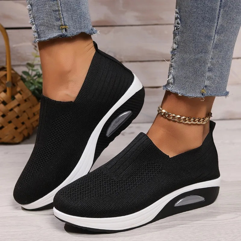 Black slip-on platform sneakers with knit mesh upper and white outsole, ankle chain.
